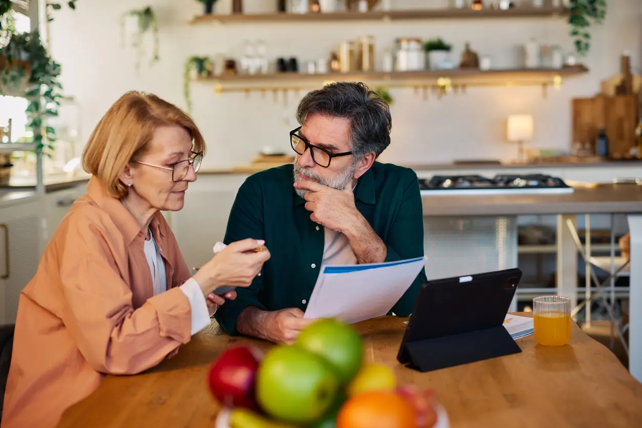 Senior couple reviewing household bills and finances at home kitchen table, thoughtful budgeting conversation Couple discussing charitable giving and finances